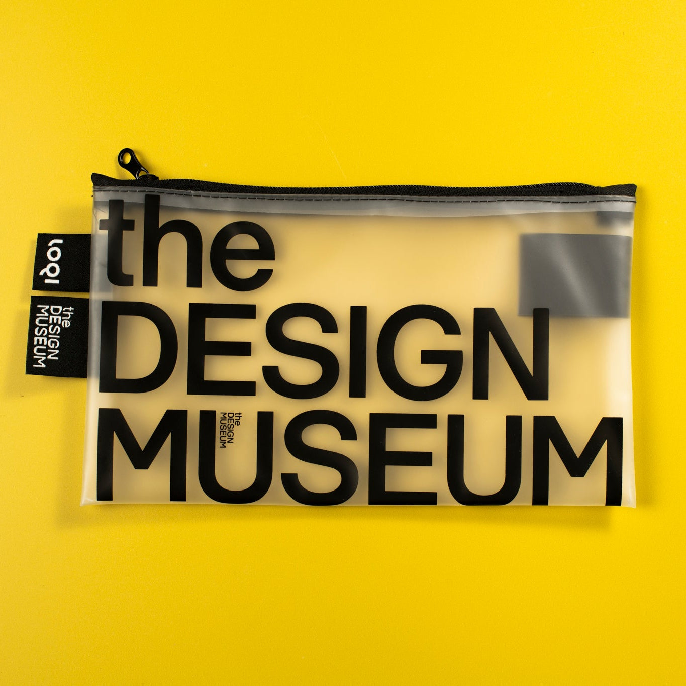 A clear zippered pouch with black text reading 'the DESIGN MUSEUM' on a bright yellow background. A black fabric tag on the left side shows the brand 'tooy'.