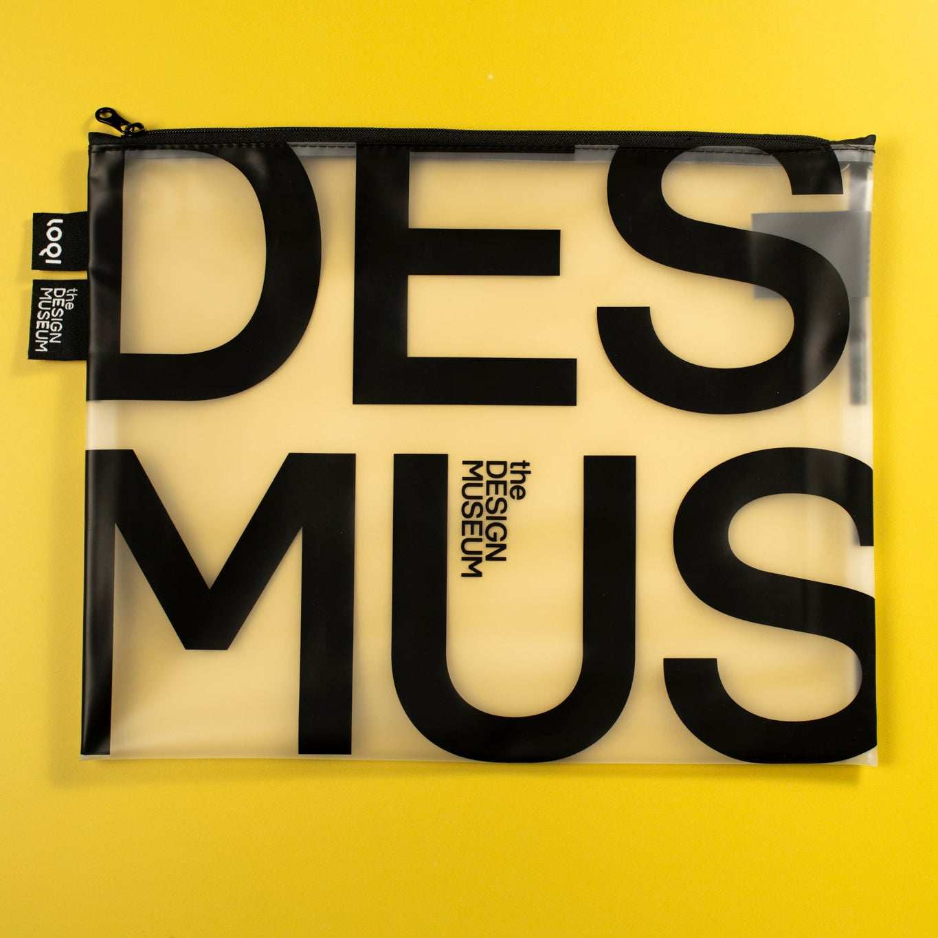 A clear zippered pouch with bold black typography reading 'DES MUS' on a bright yellow background. A small black fabric tag on the left shows the brand 'logos design studio'.