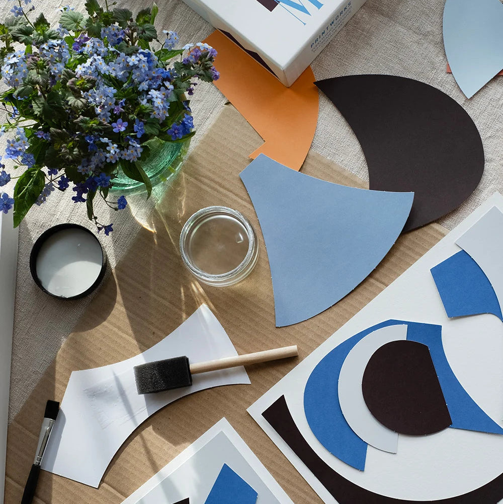 An overhead styled flat-lay of abstract art print sheets with geometric shapes on a light wood table alongside a paintbrush, small paint palette, glass jar, and a bouquet of blue forget-me-nots.