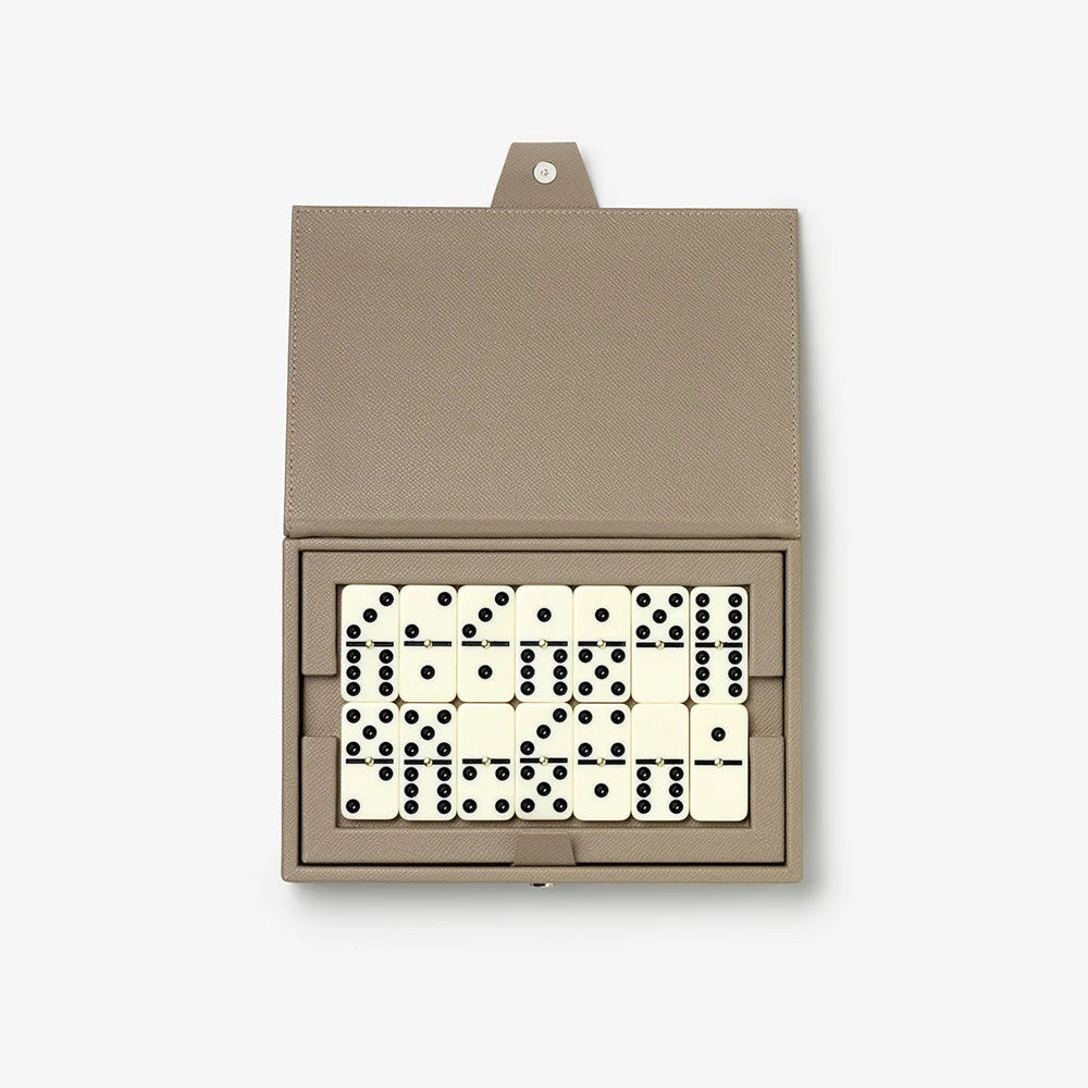 An open rectangular domino box in taupe leather, displaying a set of cream and black domino tiles arranged inside, photographed on a clean white background.