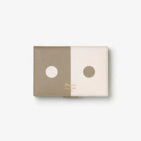 A minimalist leather domino set case featuring a split design of taupe and cream with circular cutouts, shown on a light gray background.