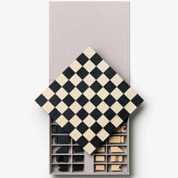A minimalist wooden chess set with a black and cream checkered board, shown partially open to reveal black and natural wood pieces in storage compartments, set against a clean white background.