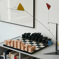 A modern minimalist wooden chess board on a shelf, with a framed print on a white wall.