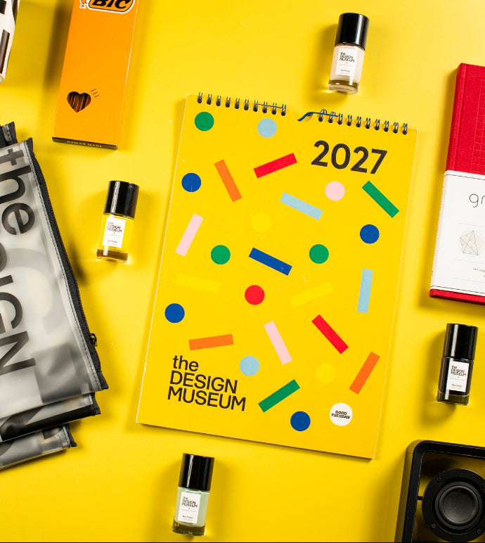 A styled flat-lay of various design and lifestyle products on a bright yellow background, including a yellow 2027 spiral notebook with colorful confetti graphics and 'the design museum' text, a black and white mesh clutch bag with bold lettering, a white mug with black stripes, and several small amber glass bottles.