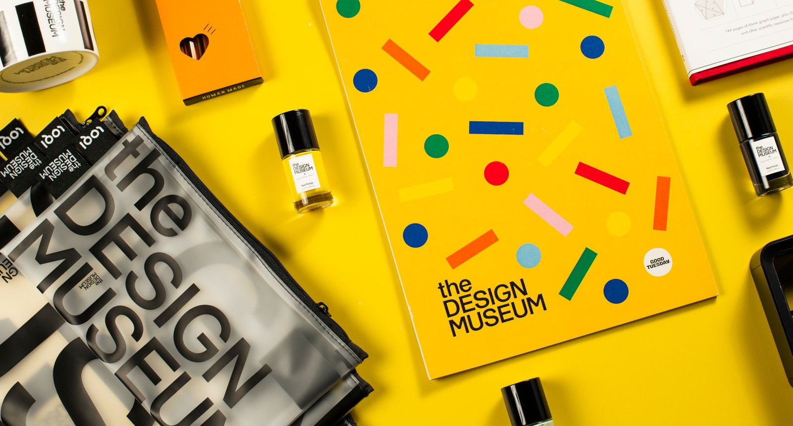 A styled flat-lay featuring a bright yellow 2027 spiral-bound notebook with colorful confetti graphics and 'the DESIGN MUSEUM' printed on the cover, surrounded by various branded items including BIC, The Design Museum bags, a mug, and a red notebook on a solid yellow background.