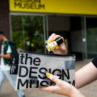 A person holds a clear zip pouch printed with 'the DESIGN MUSEUM' in large black letters, with a small yellow bottle in their other hand, in front of the Design Museum entrance.
