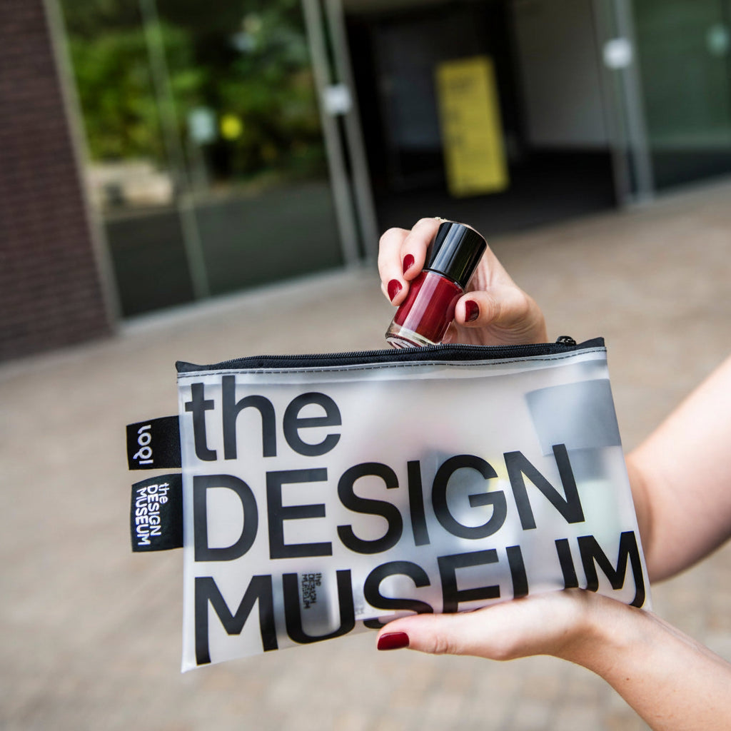 A person holds a clear zip pouch printed with large black 'the DESIGN MUSEUM' text in front of a real building entrance with a yellow sign. A hand and part of the body are visible, so this is Lifestyle rather than Product/Packaging.