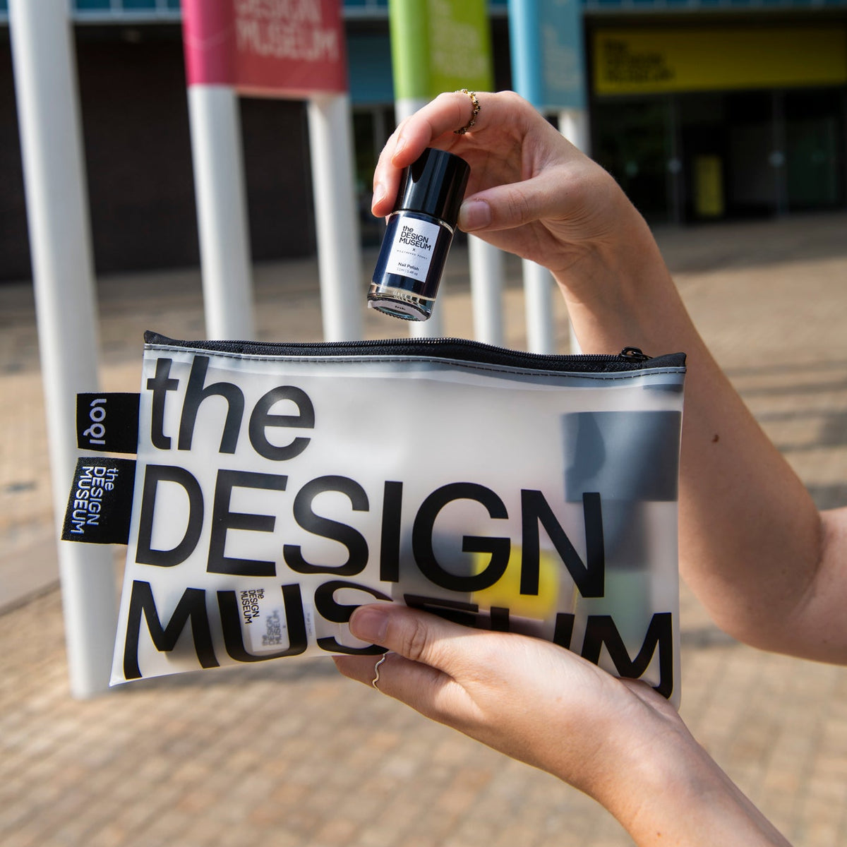 A person holds a clear zip pouch printed with large black text reading 'the DESIGN MUSEUM' in an outdoor setting, while another hand holds a small black bottle above the pouch.