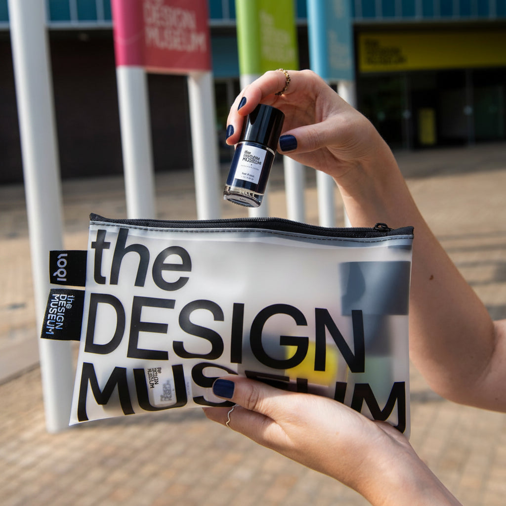 A person holds a clear zip pouch printed with large black text reading 'the DESIGN MUSEUM' in an outdoor setting, while another hand holds a small black bottle above the pouch.