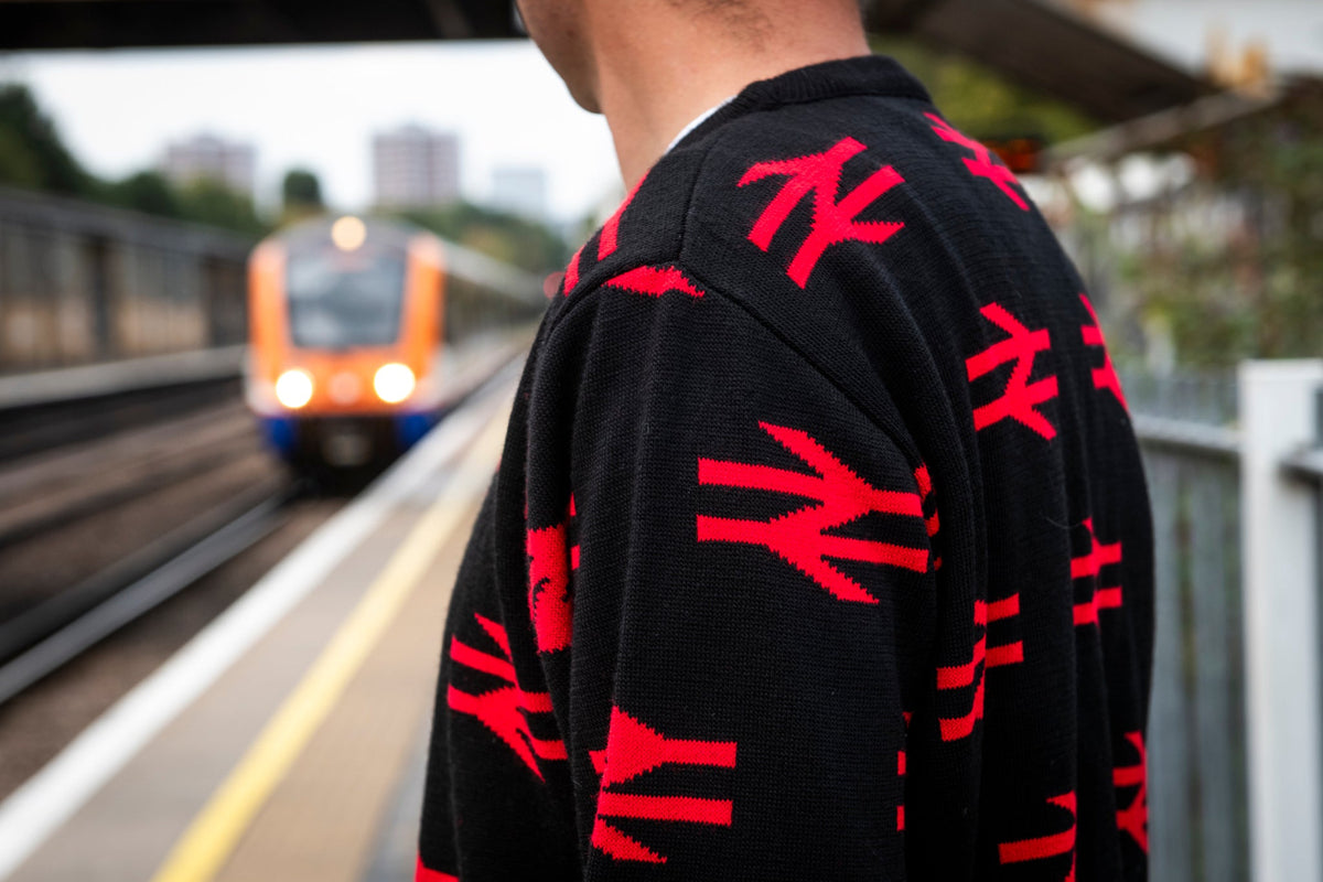 A person standing on a train station platform wearing a black knit sweater with a repeating red railway line logo pattern, with an orange train blurred in the background.