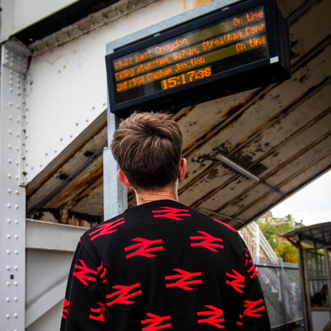 A man viewed from behind wearing a black crewneck sweatshirt with a repeating red railway logo pattern, standing at a train station platform under a digital departure board.