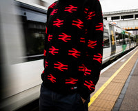 A person standing on a train station platform wearing a black sweater covered in a repeating red British Rail logo pattern, with a moving train and platform visible in the background.