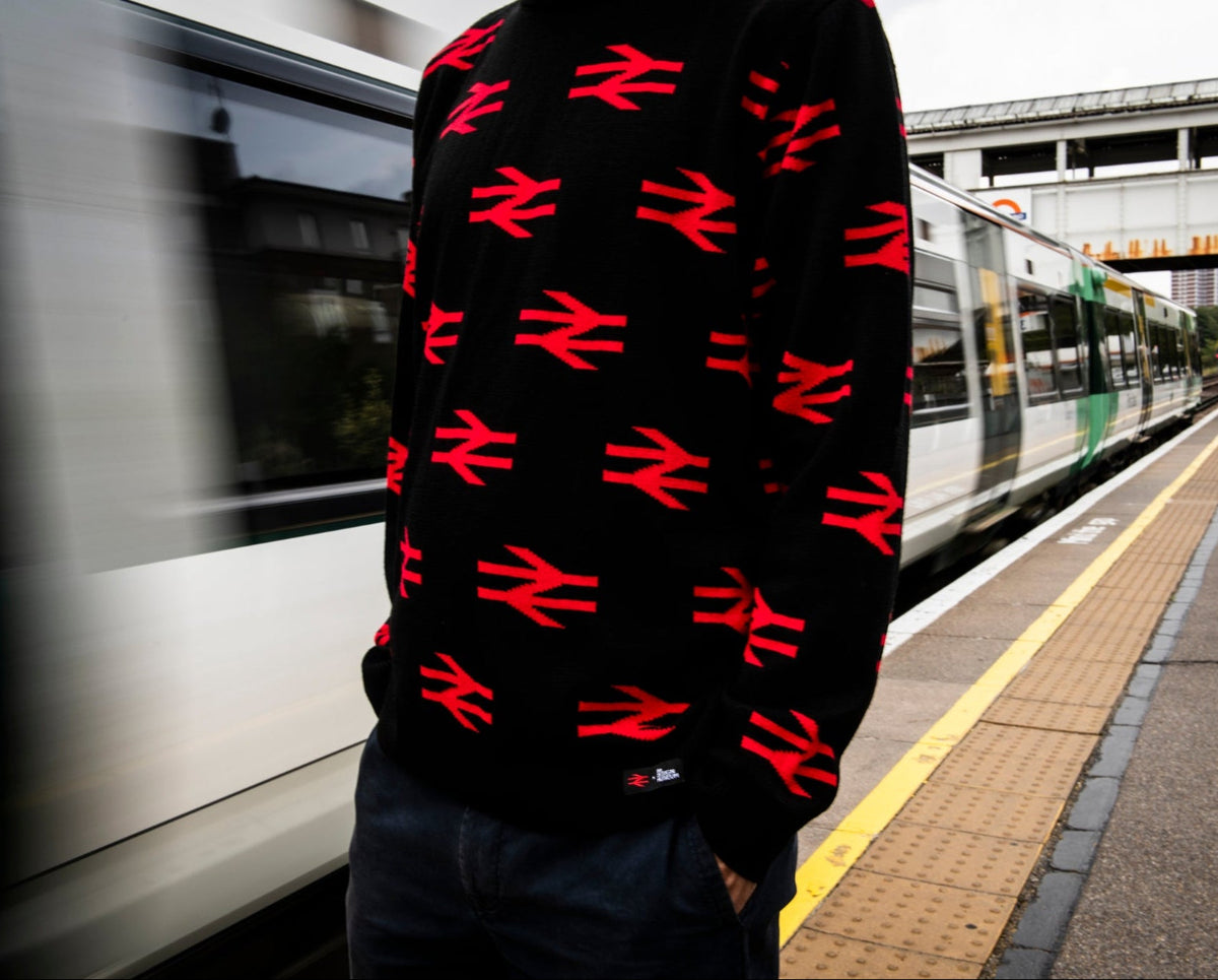 A person standing on a train station platform wearing a black sweater covered in a repeating red British Rail logo pattern, with a moving train and platform visible in the background.