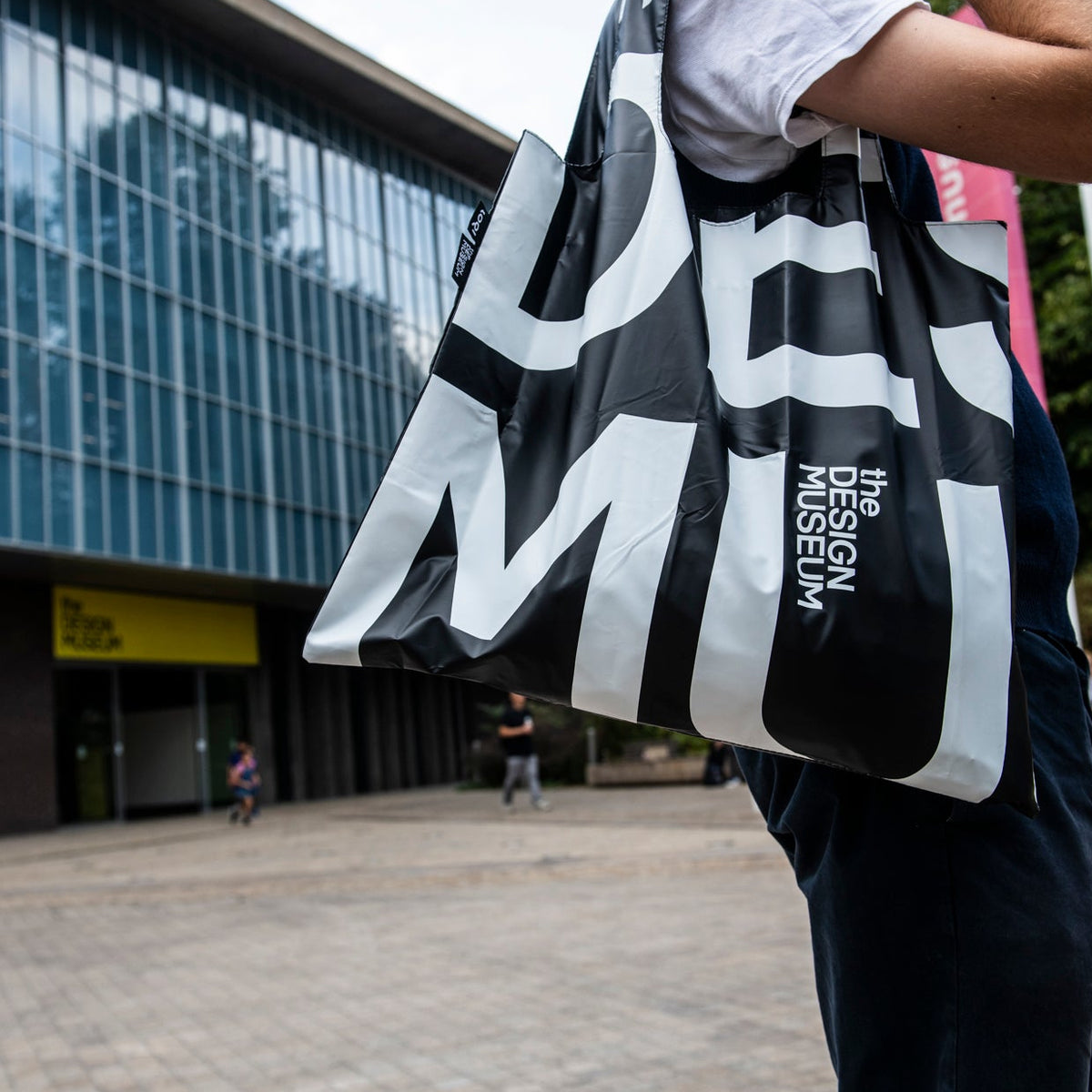 A person outdoors carrying a large black tote bag with bold white graphic lettering and vertical text reading 'DESIGN NOISE MEDIA'. The background shows a modern glass building and paved plaza.