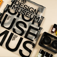 A styled flat-lay of various design and beauty products on a warm beige background, featuring a prominent clear zippered pouch with bold black typography reading 'the DESIGN MUSEUM USE MUS'. Surrounding items include an orange YBC box, a striped container, small cosmetic bottles, and a black organizer tray.