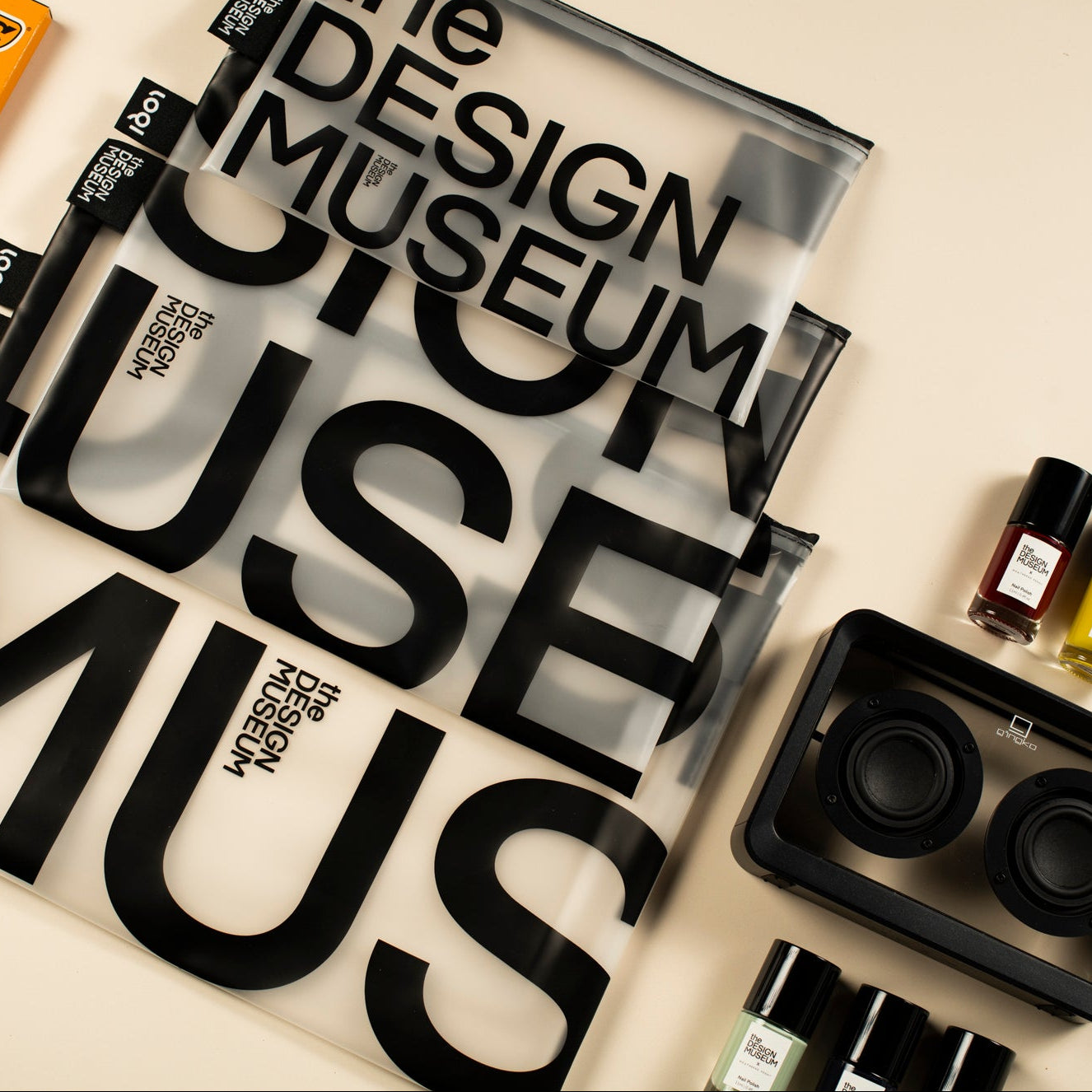 A styled flat-lay of various design and beauty products on a warm beige background, featuring a prominent clear zippered pouch with bold black typography reading 'the DESIGN MUSEUM USE MUS'. Surrounding items include an orange YBC box, a striped container, small cosmetic bottles, and a black organizer tray.