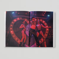 Open page of Queer London book featuring a photograph of three people dancing on stage with red lighting in front of an illuminated heart.