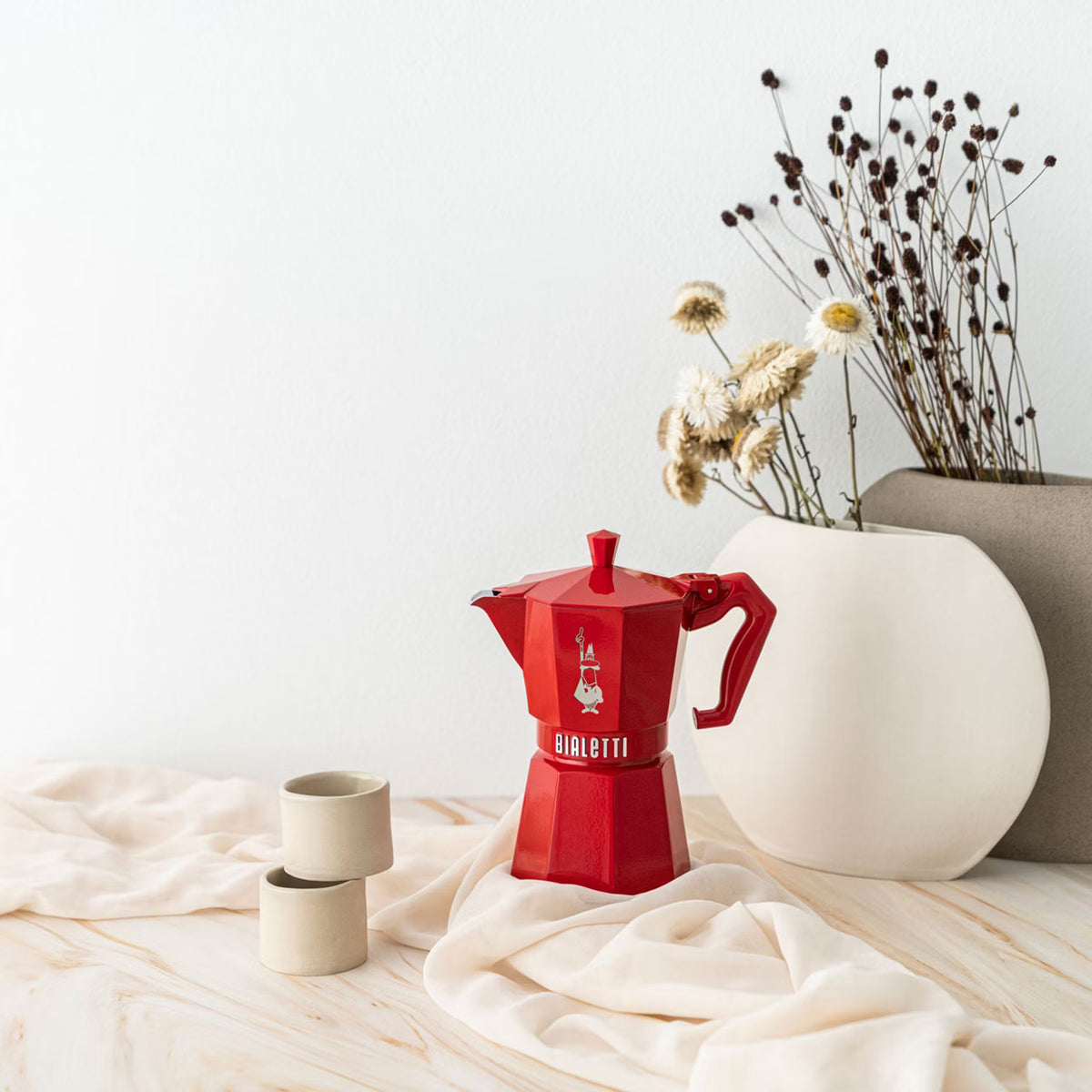 A red Bialetti Moka pot displayed on a white table cloth next to some round vases.