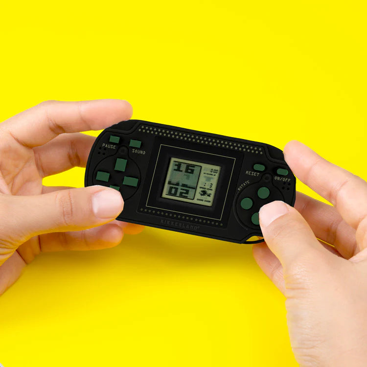 A pair of hands holding a black retro arcade game with a digital screen, pressing green buttons on a yellow backdrop.