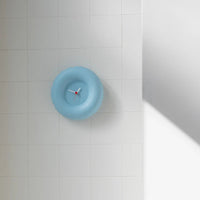 A light blue minimalist wall clock with white hands on a white tiled wall