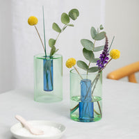 A small blue and yellow reversible vase with a clear section revealing a blue interior, displayed on a table with some floral decorations inside it.