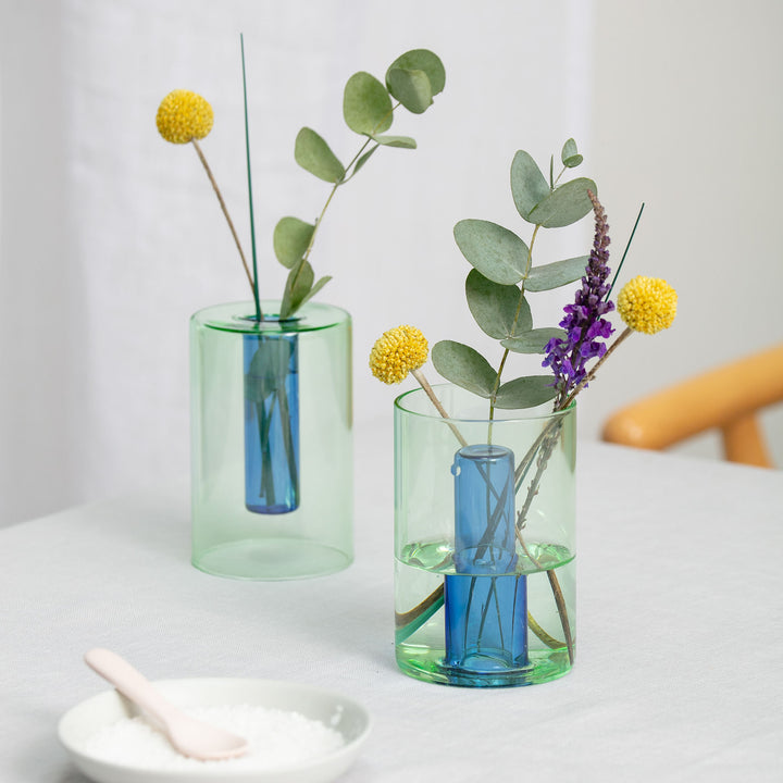 A small blue and yellow reversible vase with a clear section revealing a blue interior, displayed on a table with some floral decorations inside it.