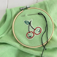 A green T-shirt embroidered with cherries with a needle and an embroidery hoop.