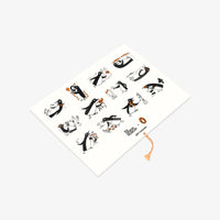 A white rectangular notebook or journal with a decorative cover featuring black and orange penguin illustrations, photographed on a light gray background.