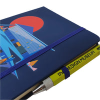 Close up image of the Design Museum Branded A5 size notebook front cover to detail its elastic bands to keep notebook closed and elastic band slot for a pen or pencil with a pen in it.
