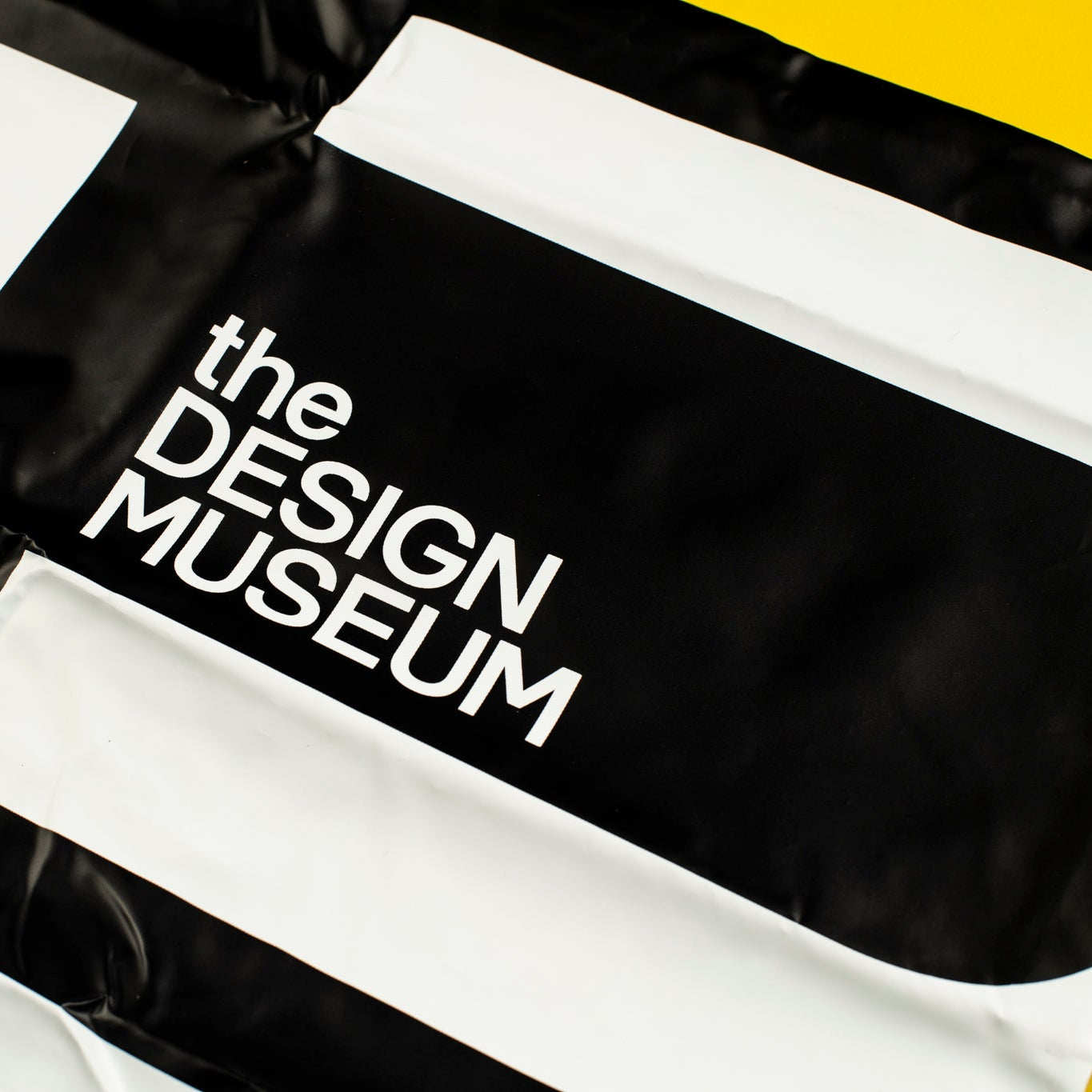 A close-up of a black and white striped flag or banner with the words 'the DESIGN MUSEUM' printed in white text on a black section. A small yellow area is visible in the top-right corner.