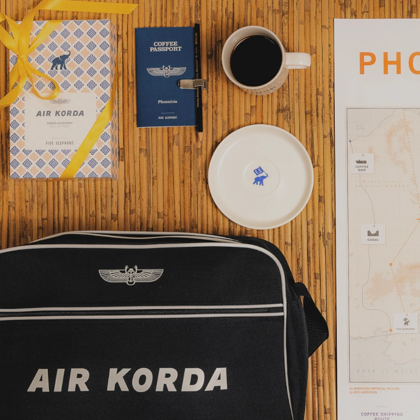 Black 'AIR KORDA' bag with a coffee cup, passport, and map on a wooden surface