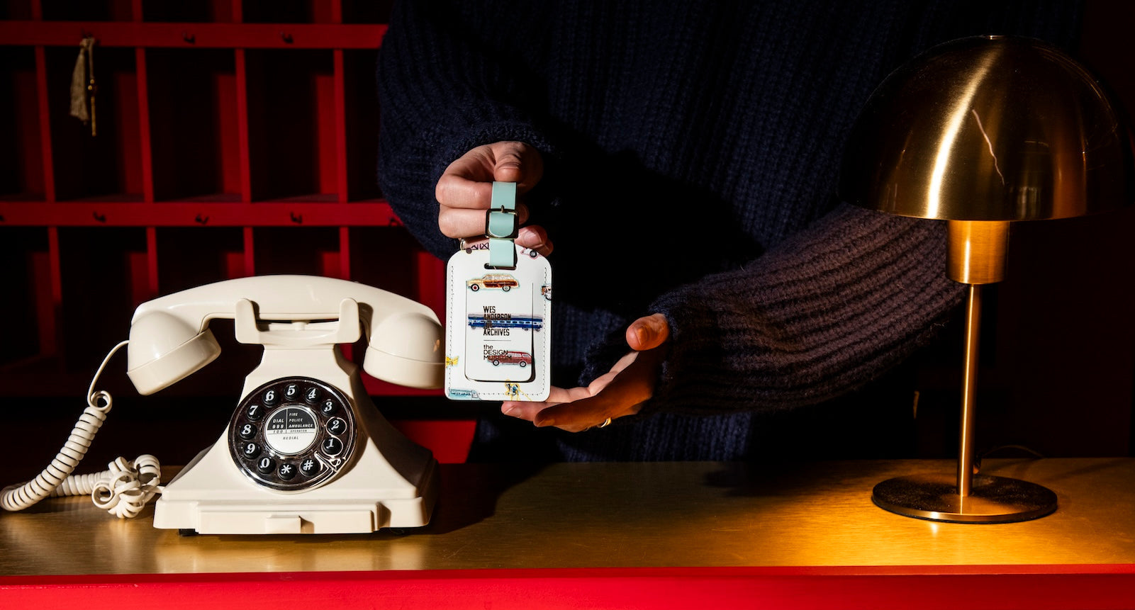 Person holding a vintage-style luggage tag next to an old-fashioned rotary phone on a desk.