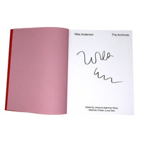 An open book or booklet showing a white page with the text 'Wes Anderson The Archives' and a black handwritten signature, alongside a large pink interior spread and a red spine edge on a white background.