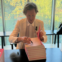 A man in a light-colored blazer and white shirt sits at a table, writing on a stack of pink-and-white books with a black pen. Large windows behind him show green autumn trees outside.