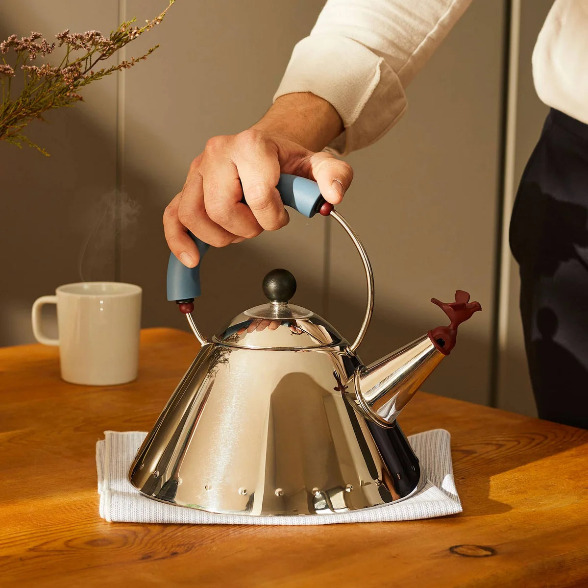 A stainless steel teapot with a blue handle grip resting on a white cloth on a wooden table, with a person's hand holding the handle.