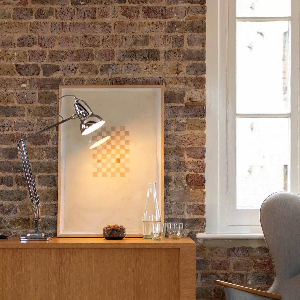 Modern interior with a wooden desk, chrome lamp, framed picture, and window on a brick wall.