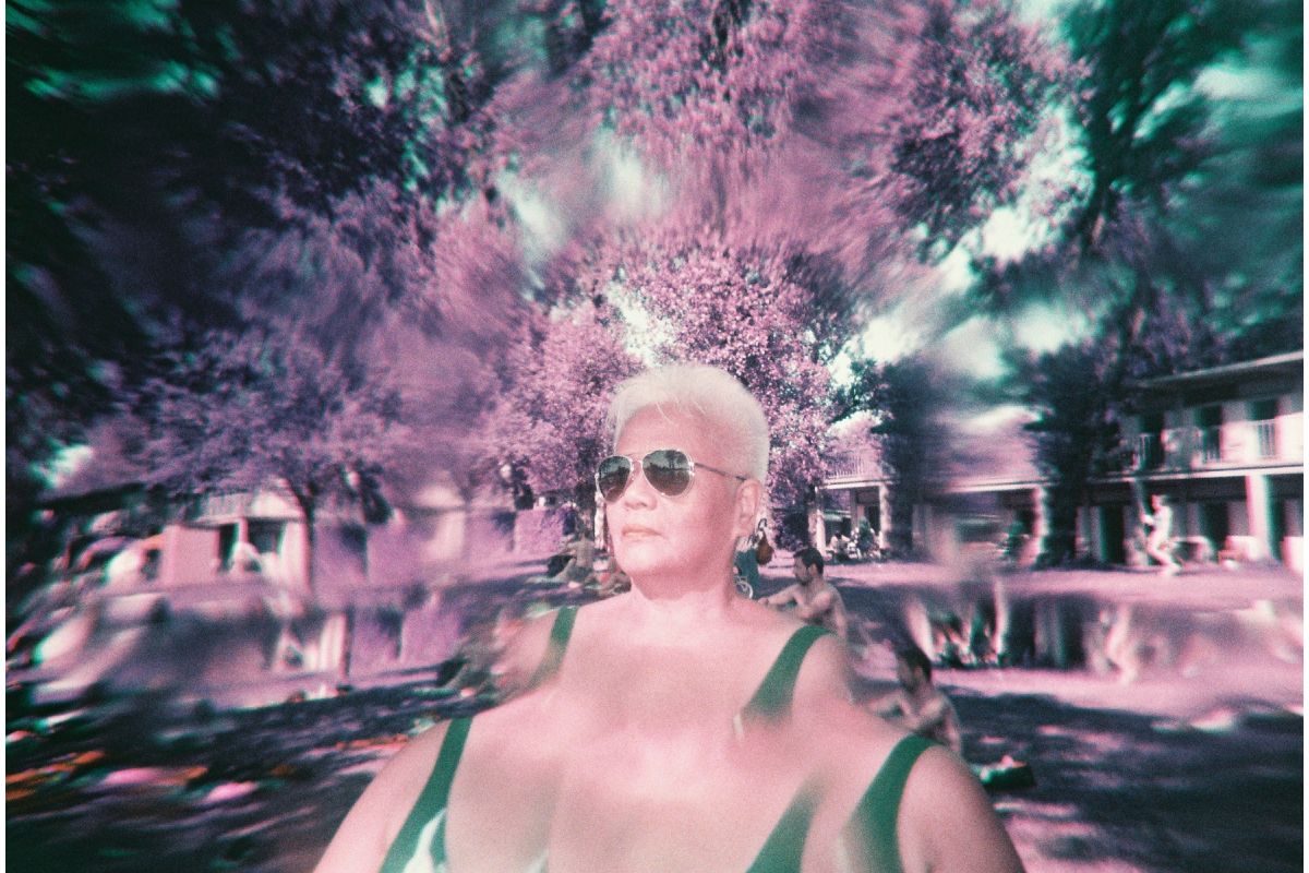 A pink distorted photo of a person with platinum blonde hair wearing sunglasses and a swimsuit in a pool setting.