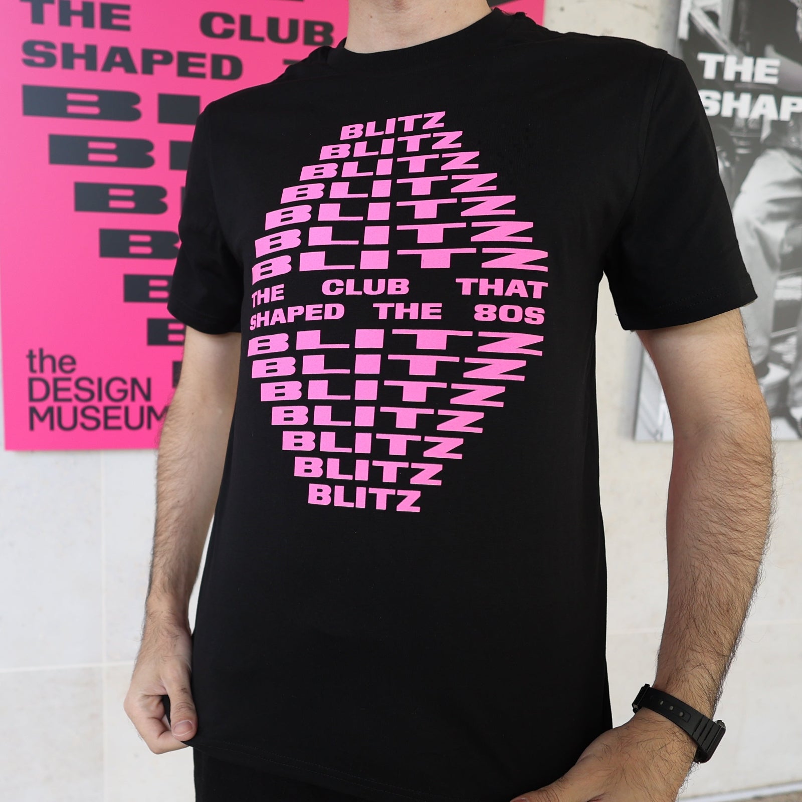 Person wearing a black t-shirt with pink text in front of a pink poster.