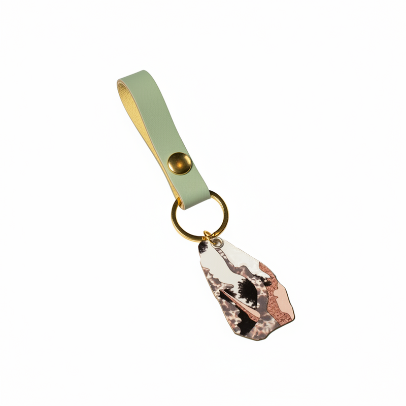 A leather luggage tag with a mint green strap, gold-tone metal hardware, and a printed acrylic charm featuring a black and white cat with pink accents.