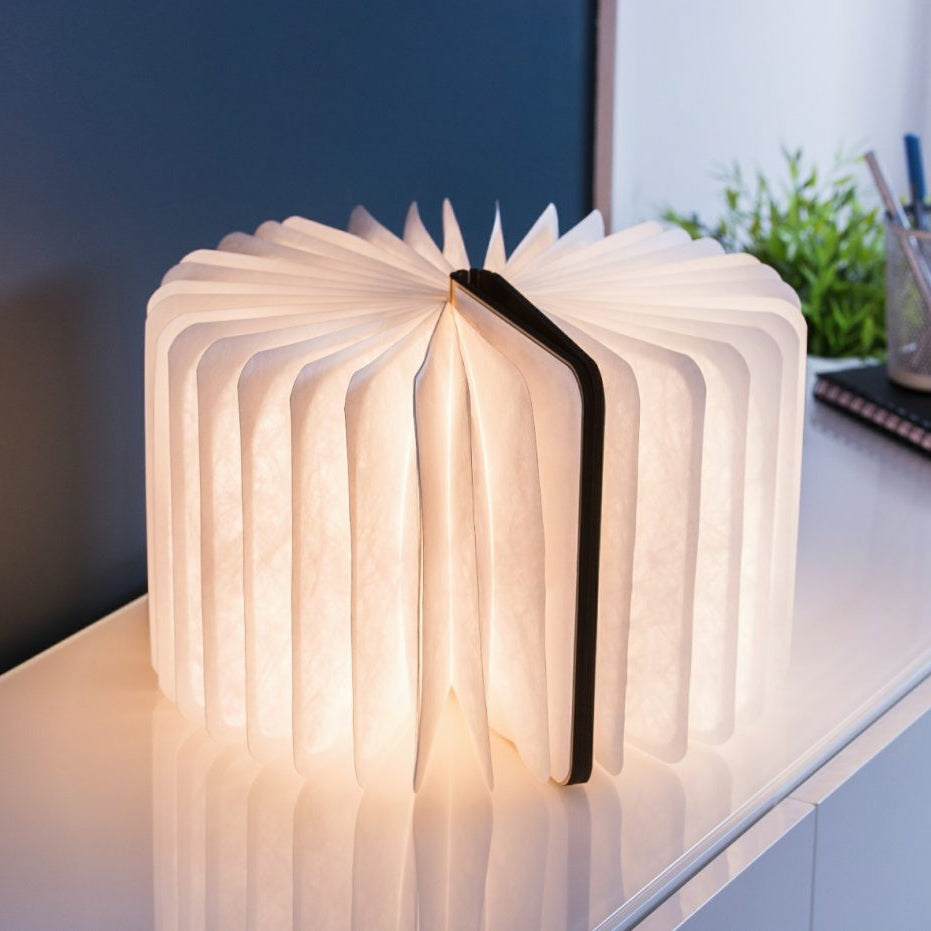 A book-shaped folding table lamp glowing with warm light on a white desk, with a dark navy wall and office accessories in the background.