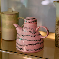 A pink and black abstract ceramic coffee pot on a gold shelf.