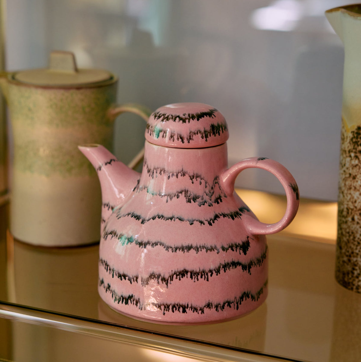 A pink and black abstract ceramic coffee pot on a gold shelf.