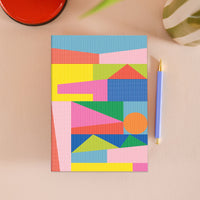 A notebook with a colorful geometric pattern made of striped paper strips, placed on a beige surface next to a blue pen.