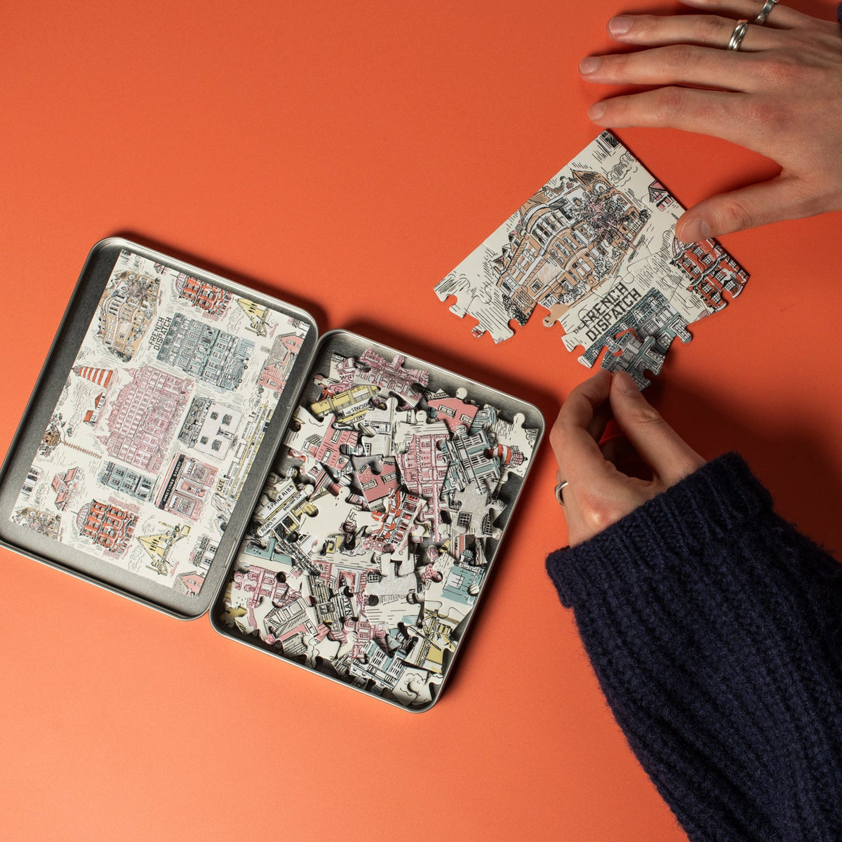 An open tin of puzzle pieces on a red surface with hands interacting with them.