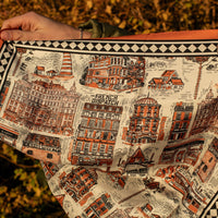 Silk scarf with architectural illustrations and an orange border in front of a bush