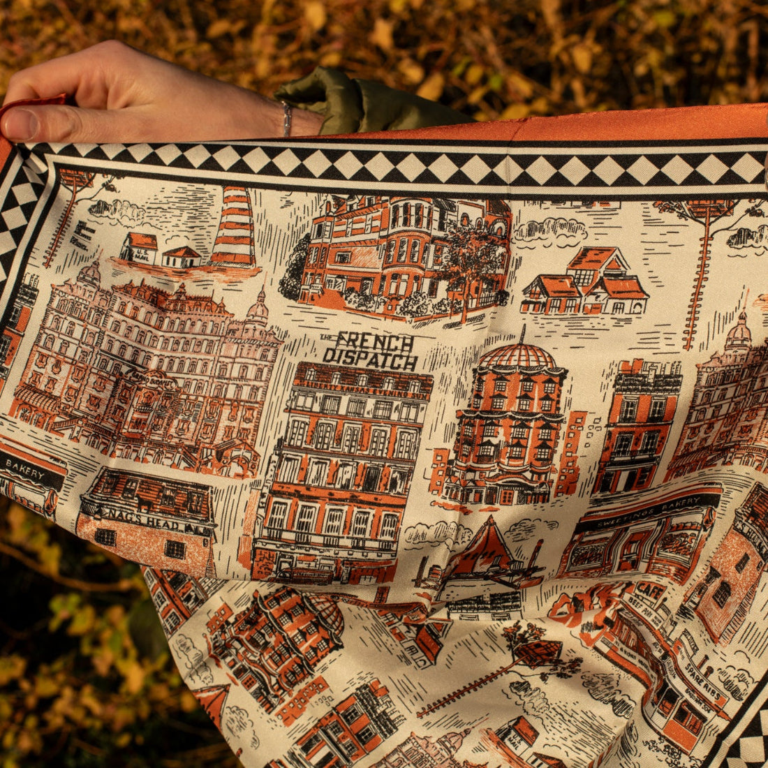 Silk scarf with architectural illustrations and an orange border in front of a bush