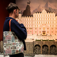 Person holding a patterned tote bag in front of a model of a large building with mountains in the background