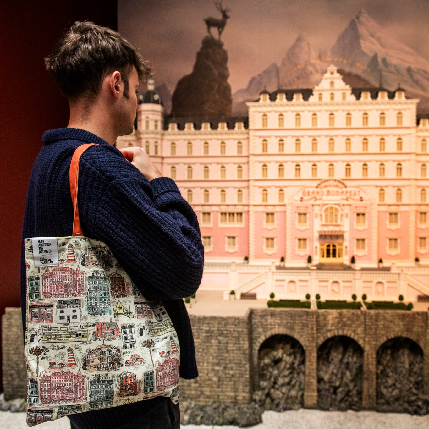 Person holding a patterned tote bag in front of a model of a large building with mountains in the background