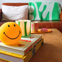 A cordless, round lamp with a yellow silicone body and a smiling face design on top of a stack of books on a wooden table.
