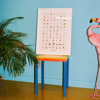 Colourful chair with a poster of a collection of chairs in front of a blue wall, next to a pink flamingo.